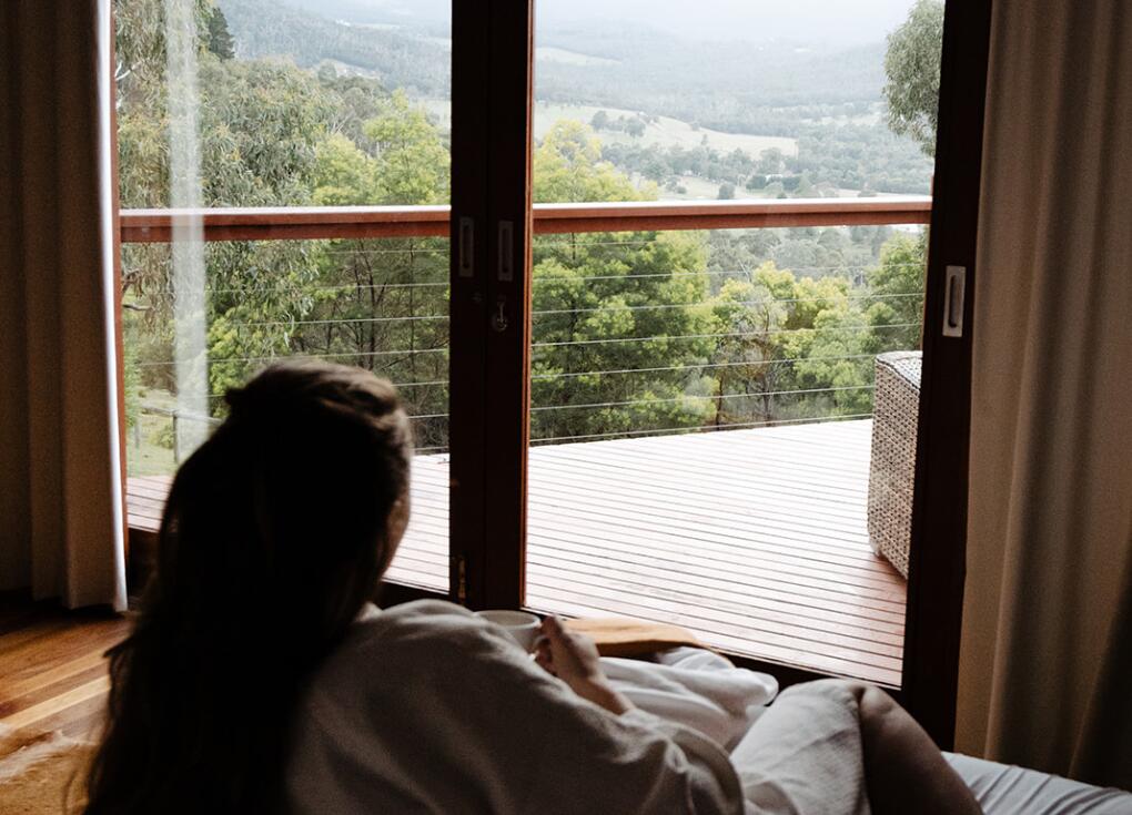 A solo woman lying on a bed drinking a coffee looking at a bush view